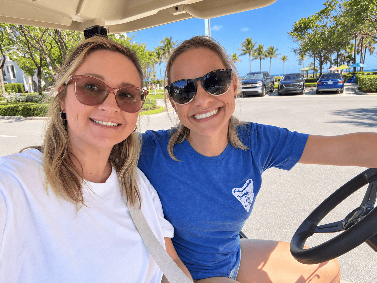 Two women smiling, one in a white shirt and sunglasses, the other in a blue shirt and sunglasses, sitting in a golf cart outdoors.