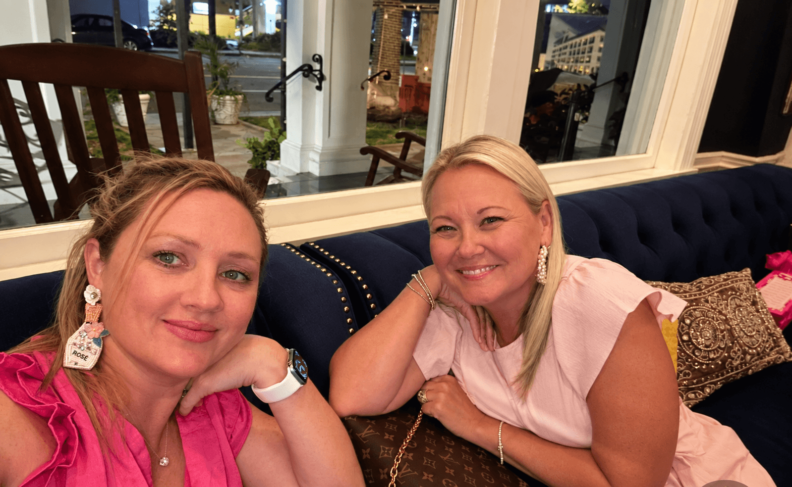 Two women sitting on a dark blue couch, smiling, in a well-lit room with large windows behind them.
