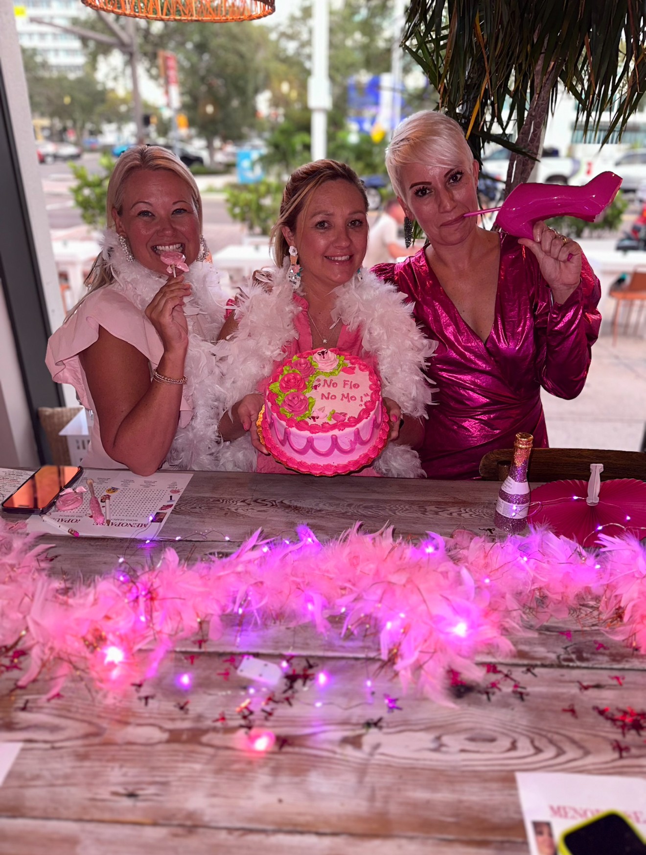Three women celebrating a birthday with a pink cake, pink feather decoration, and a pink shoe prop, smiling indoors.
