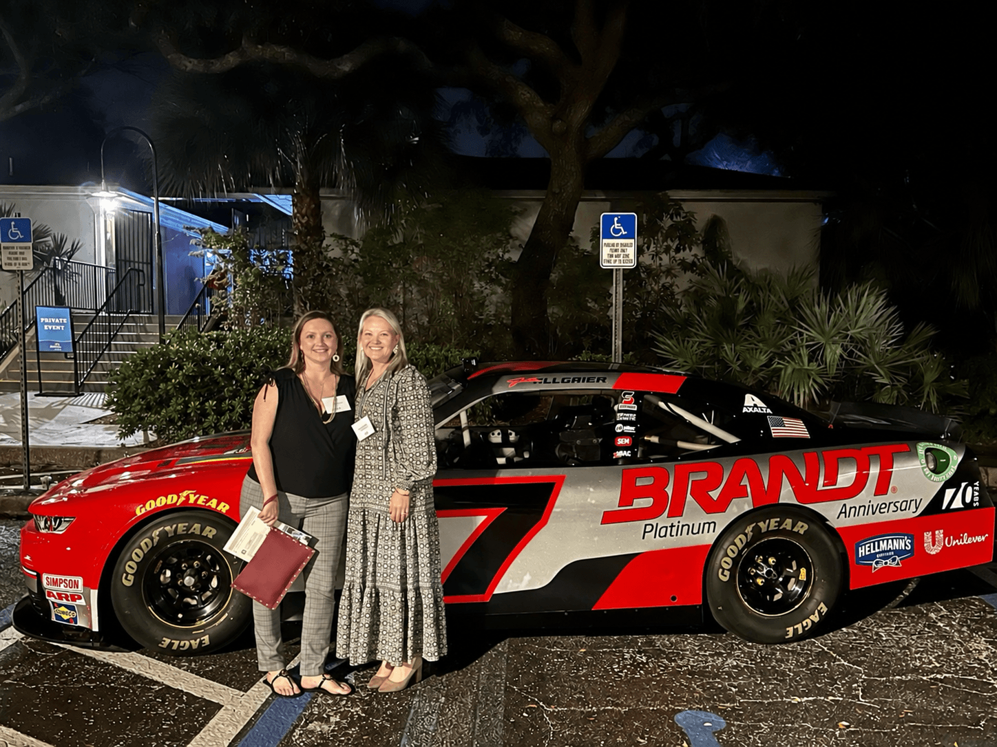 Two women stand next to a race car at night, smiling, with trees and signs in the background.