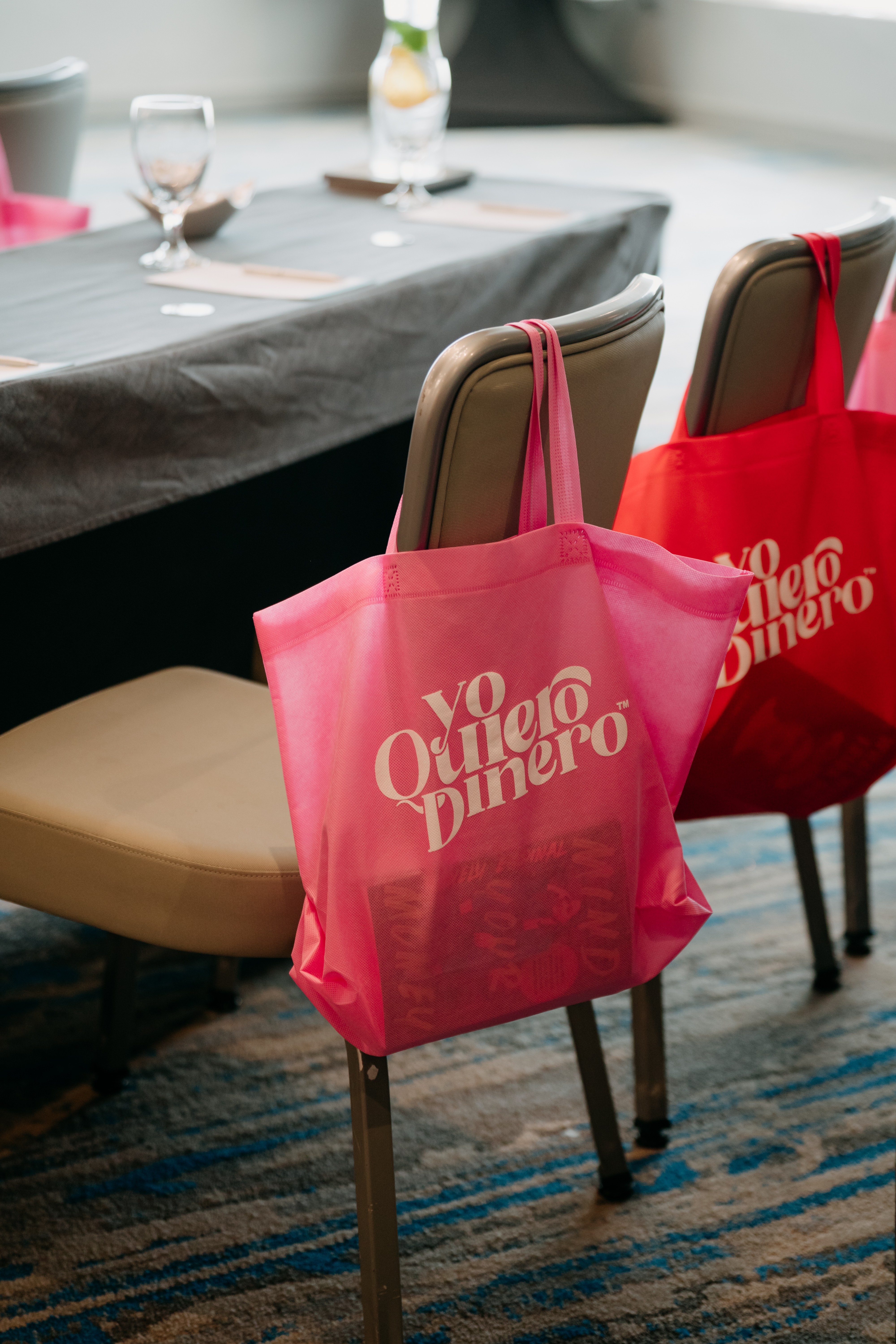 Two red tote bags with white text hang on chairs around a dining table, which has glasses and a tablecloth.