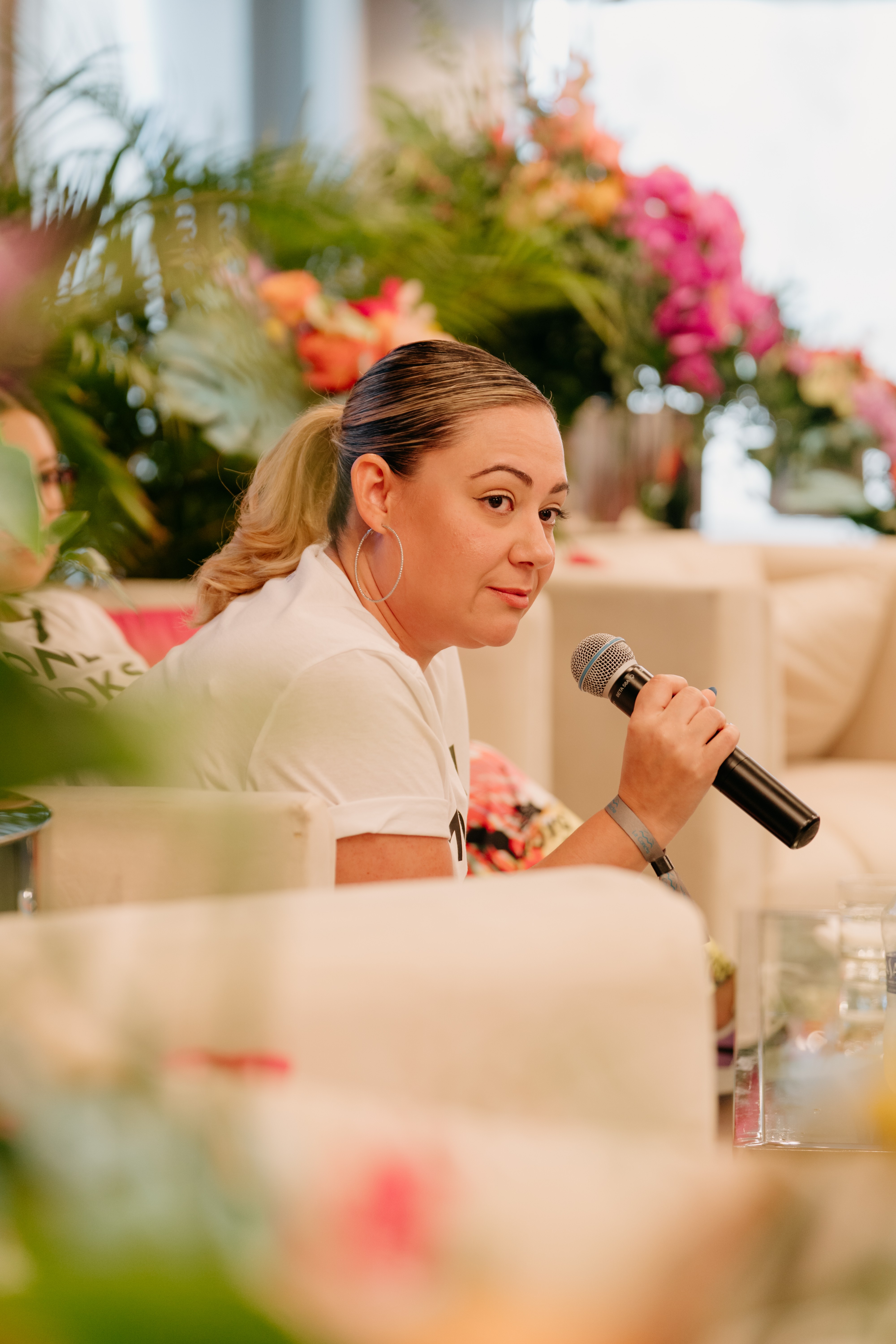 Woman holding a microphone, sitting at a table with flowers and plants in the background.