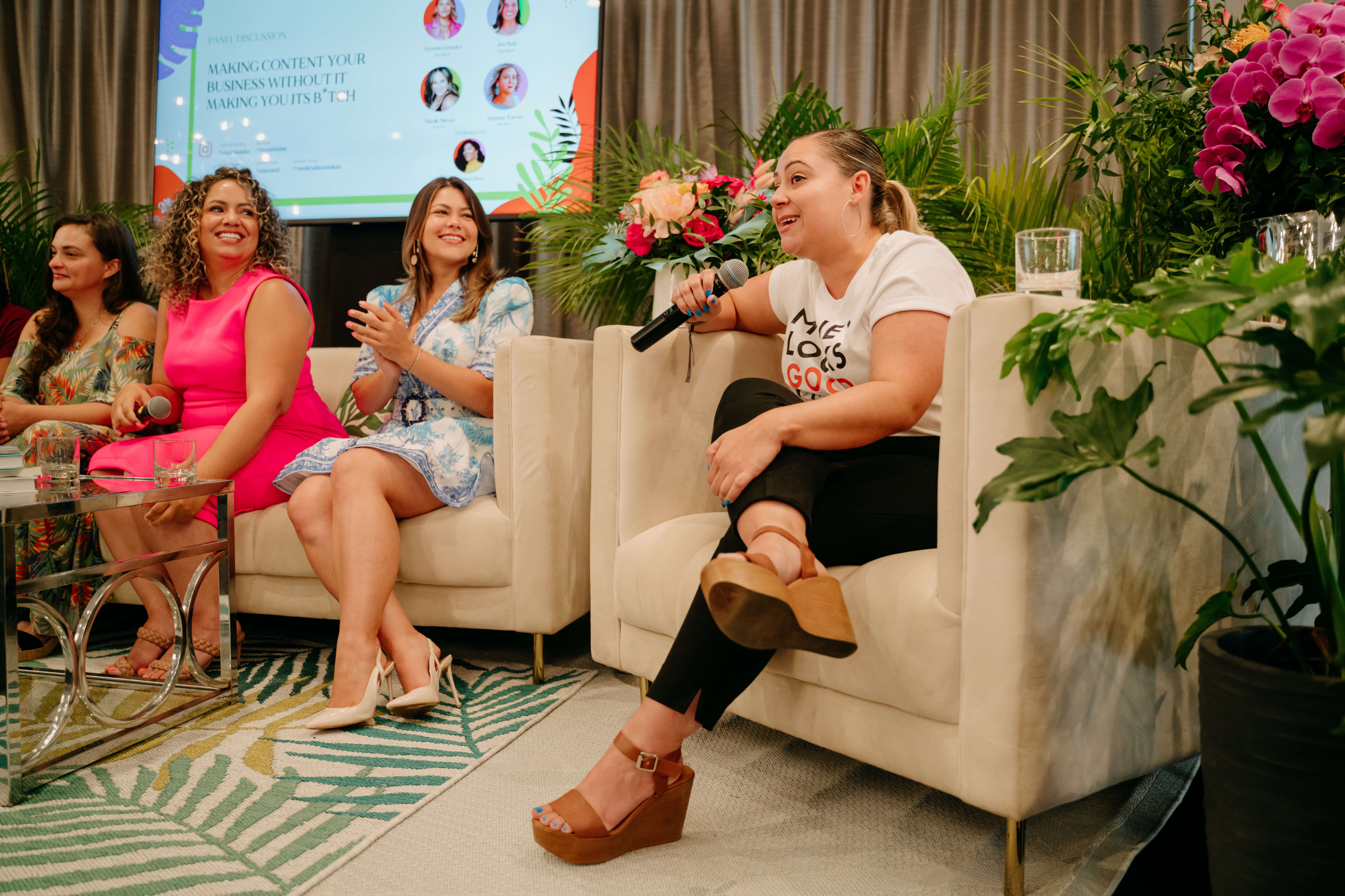 Four women sitting on a stage with flowers and a screen in the background, one woman speaking into a microphone.