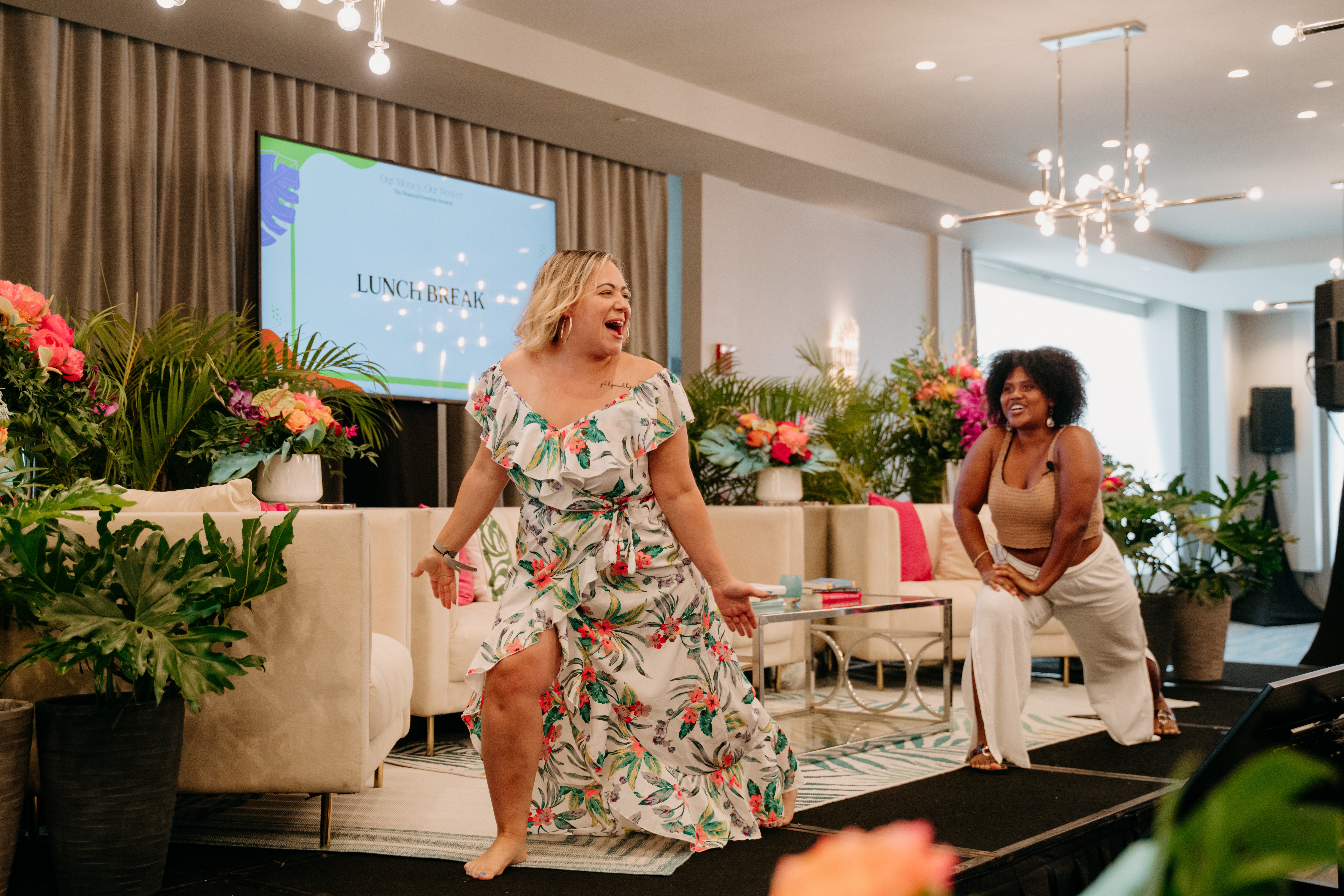 Two women laughing and dancing in a decorated room with flowers and a large screen, one in a floral dress, the other in a white outfit.