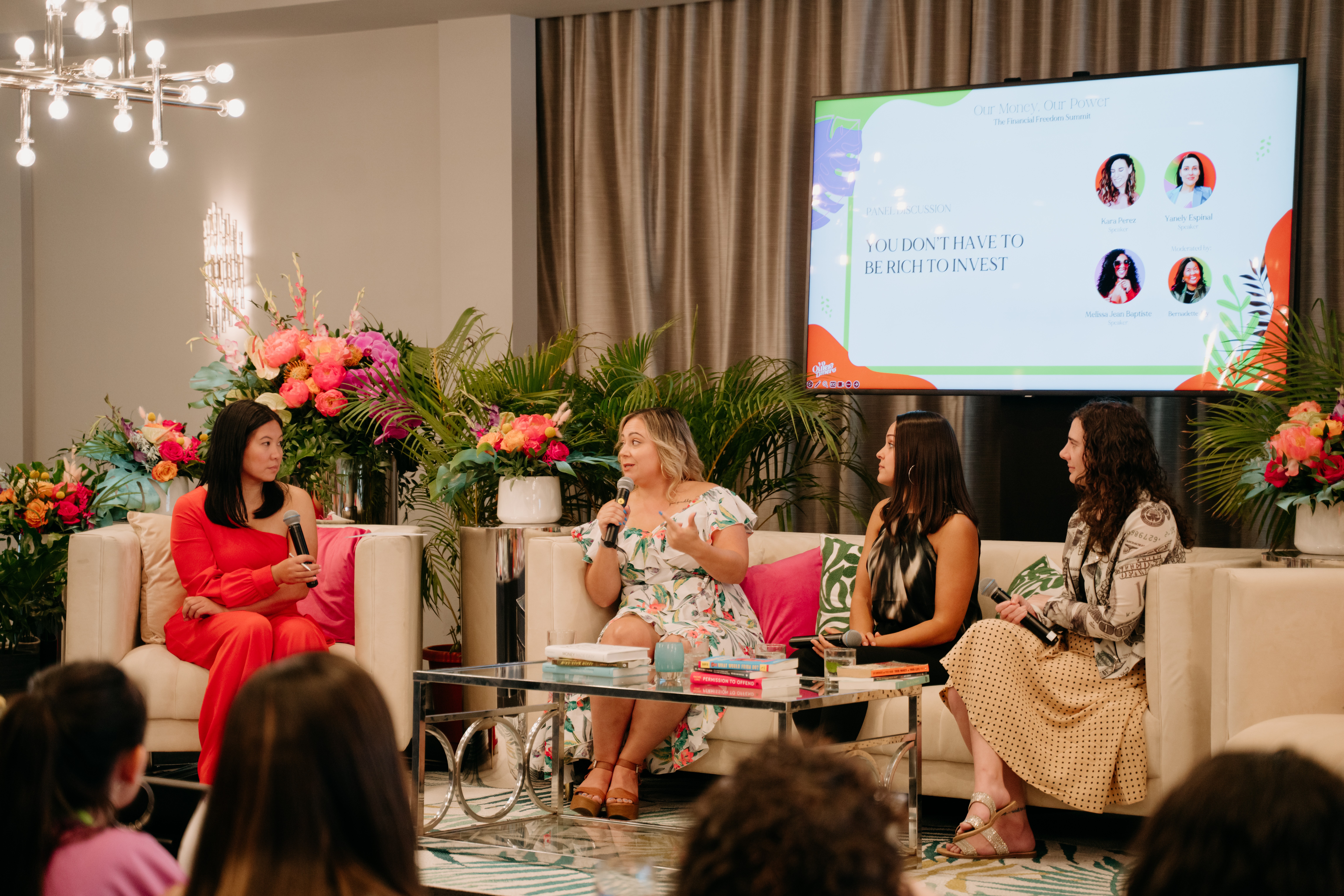 Four women seated on a stage with floral arrangements and a large screen behind them, engaging in a discussion.