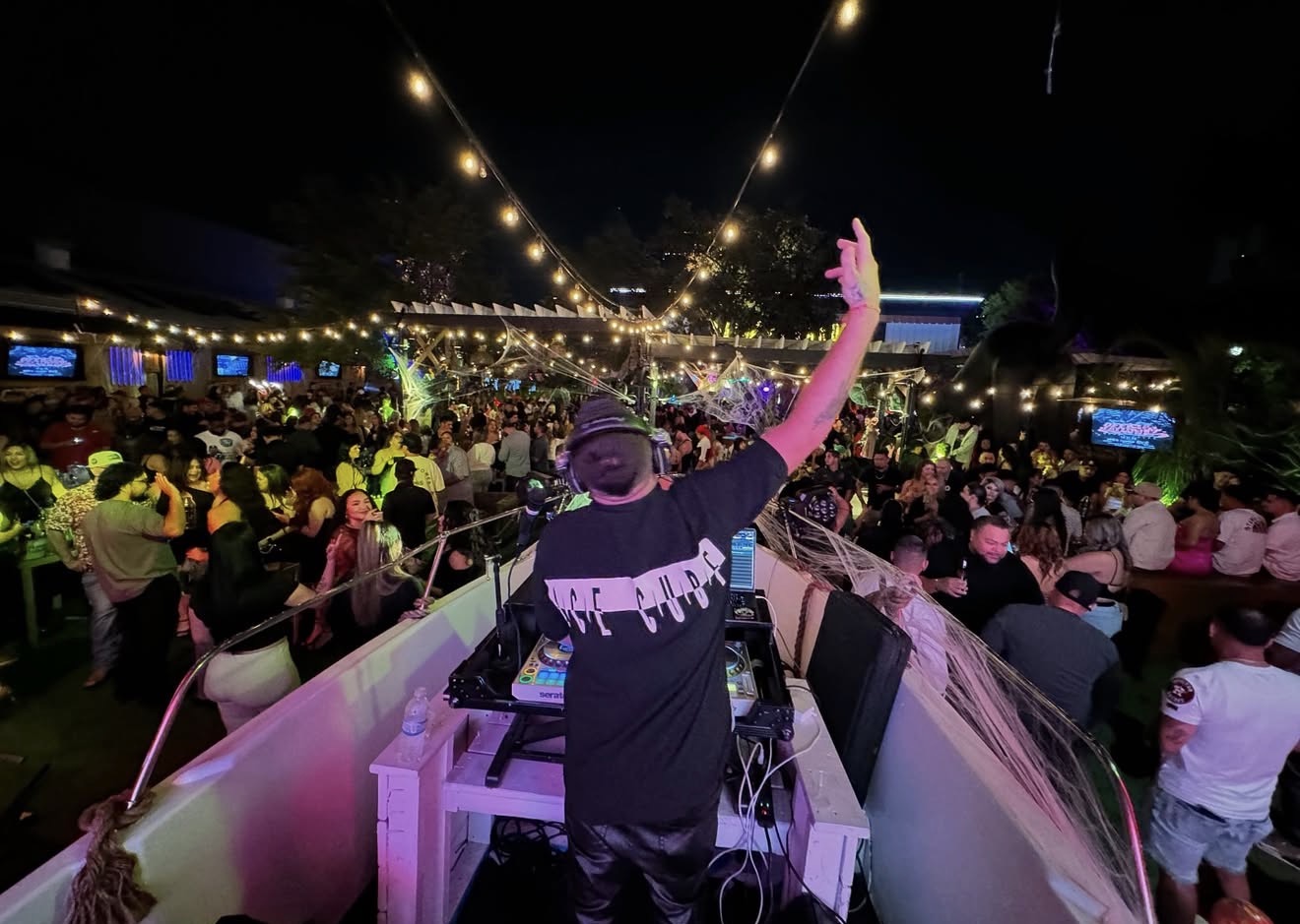 DJ performs at outdoor night event with string lights and large crowd, illuminated by string lights overhead.