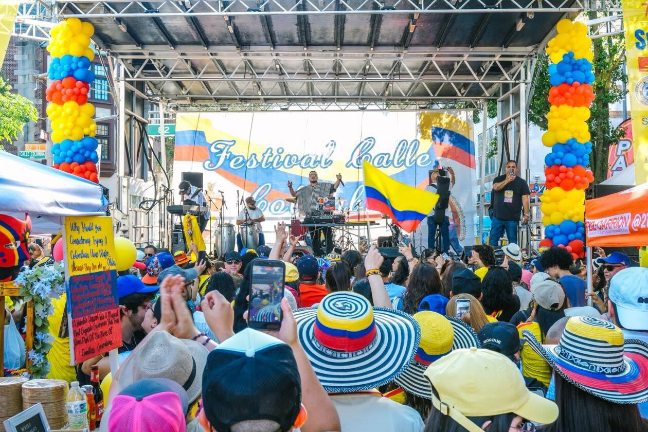Crowd watching a stage with performers, colorful balloons, and banners at a festival, many wearing hats and holding flags.