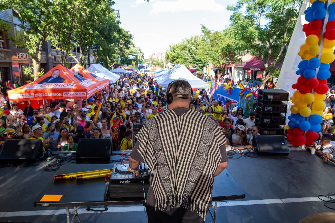 Performer on stage facing a large crowd outdoors with trees and tents, colorful balloons on the right, sunny day.