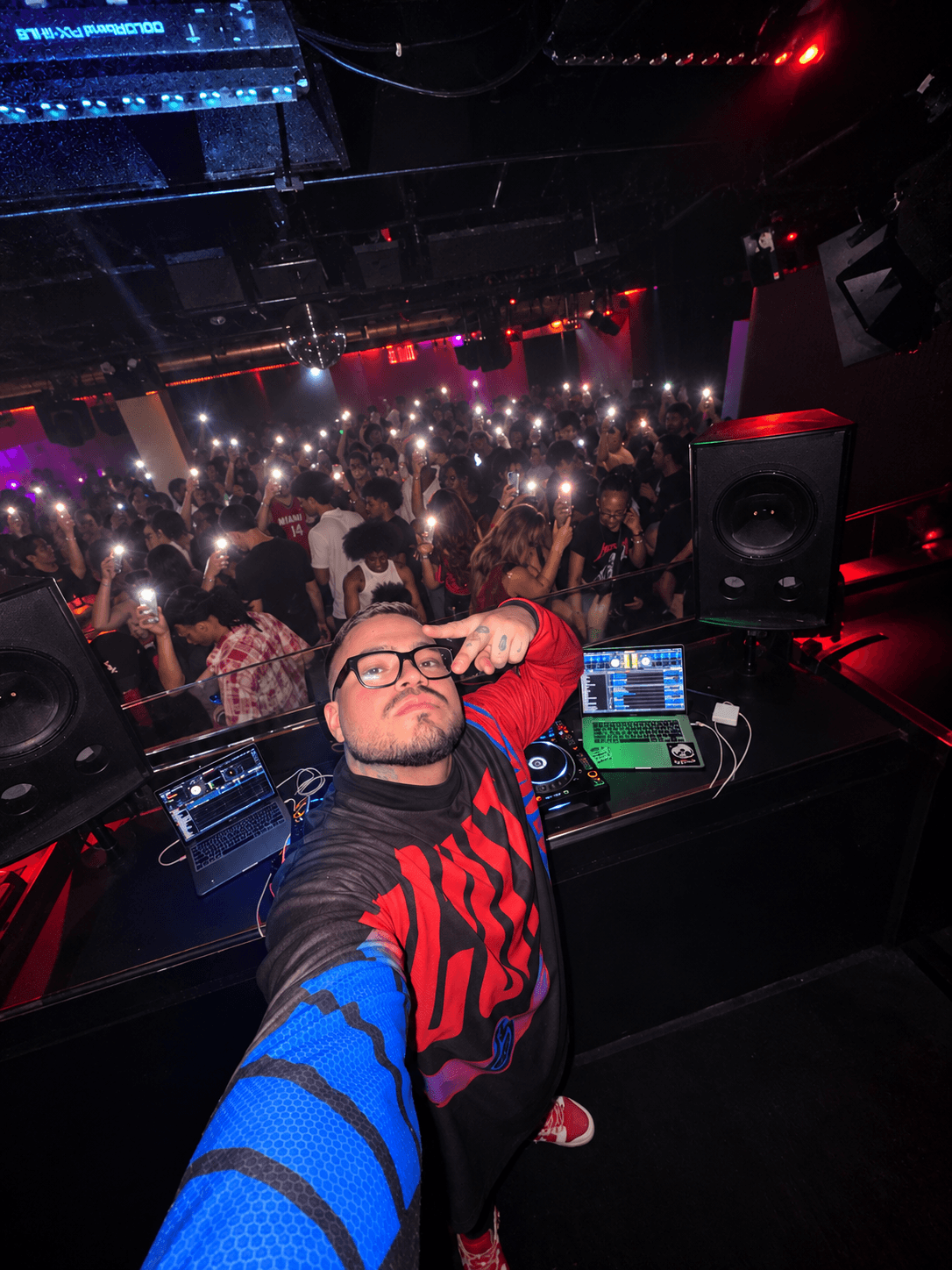 DJ taking a selfie in a nightclub with a crowd and colorful lights in the background.