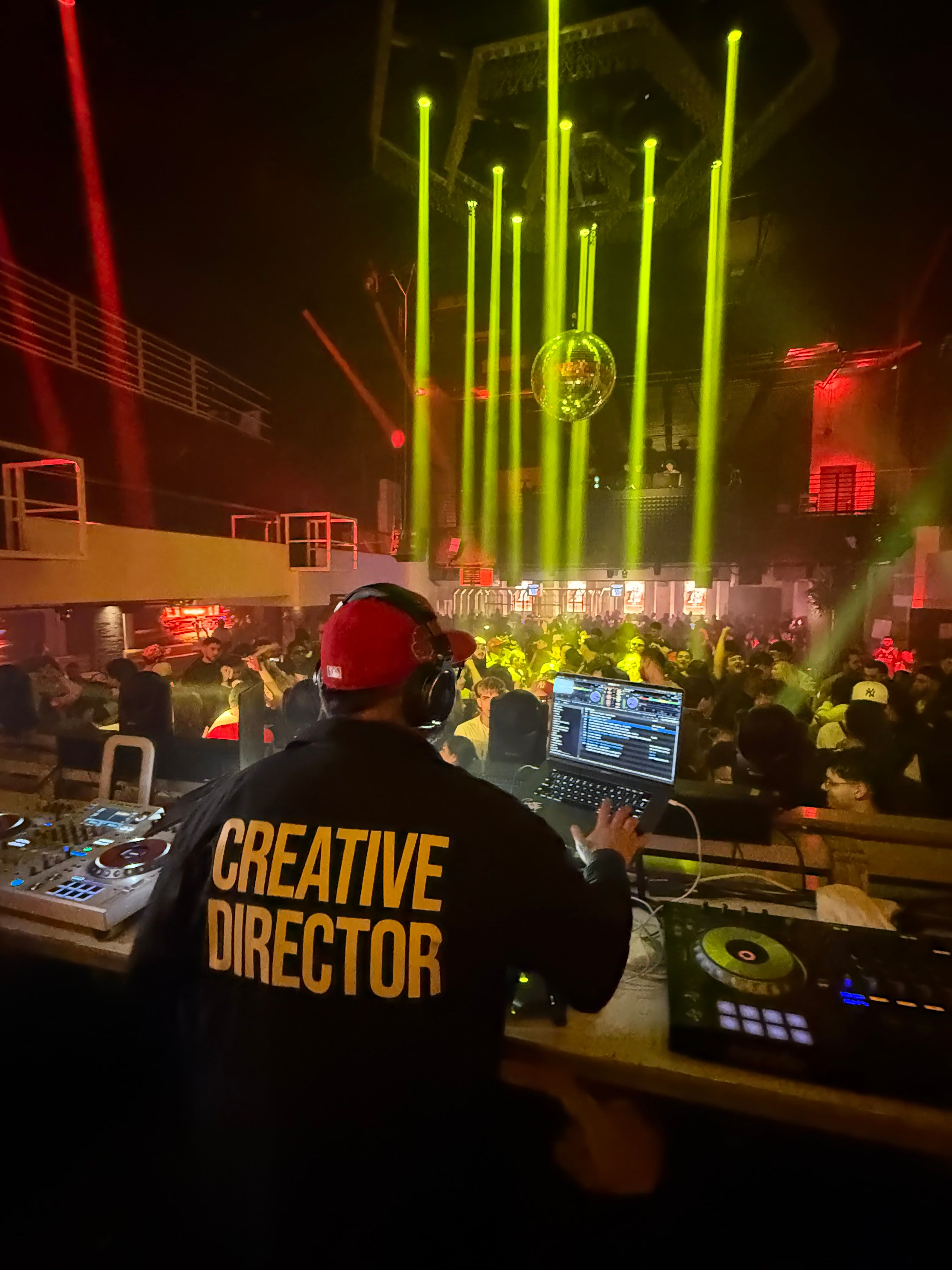 DJ performing at a nightclub with colorful lights and a crowd in the background, wearing a shirt that says 'Creative Direction'.