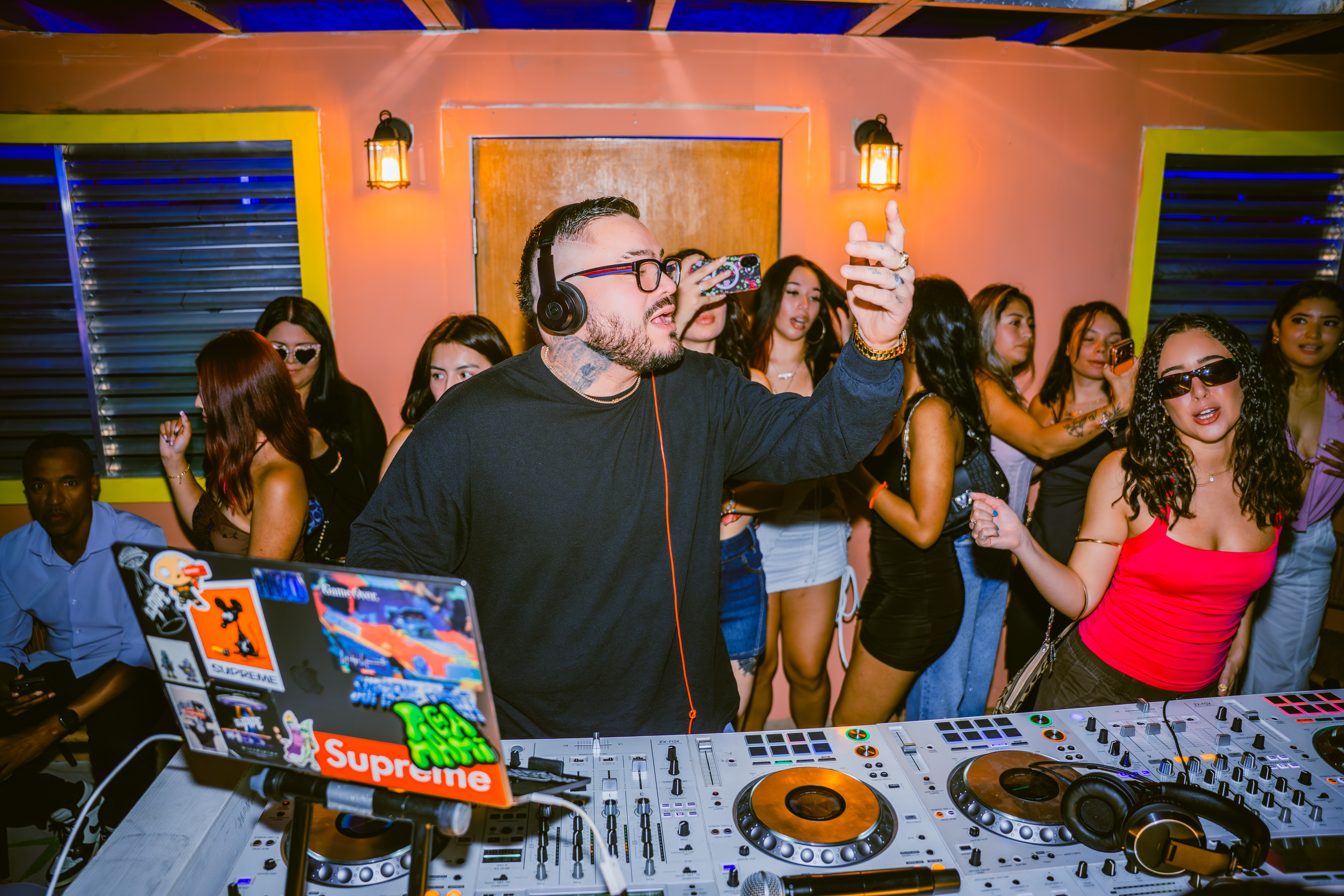 DJ with headphones and glasses raises a hand at a party with people dancing and socializing in the background.