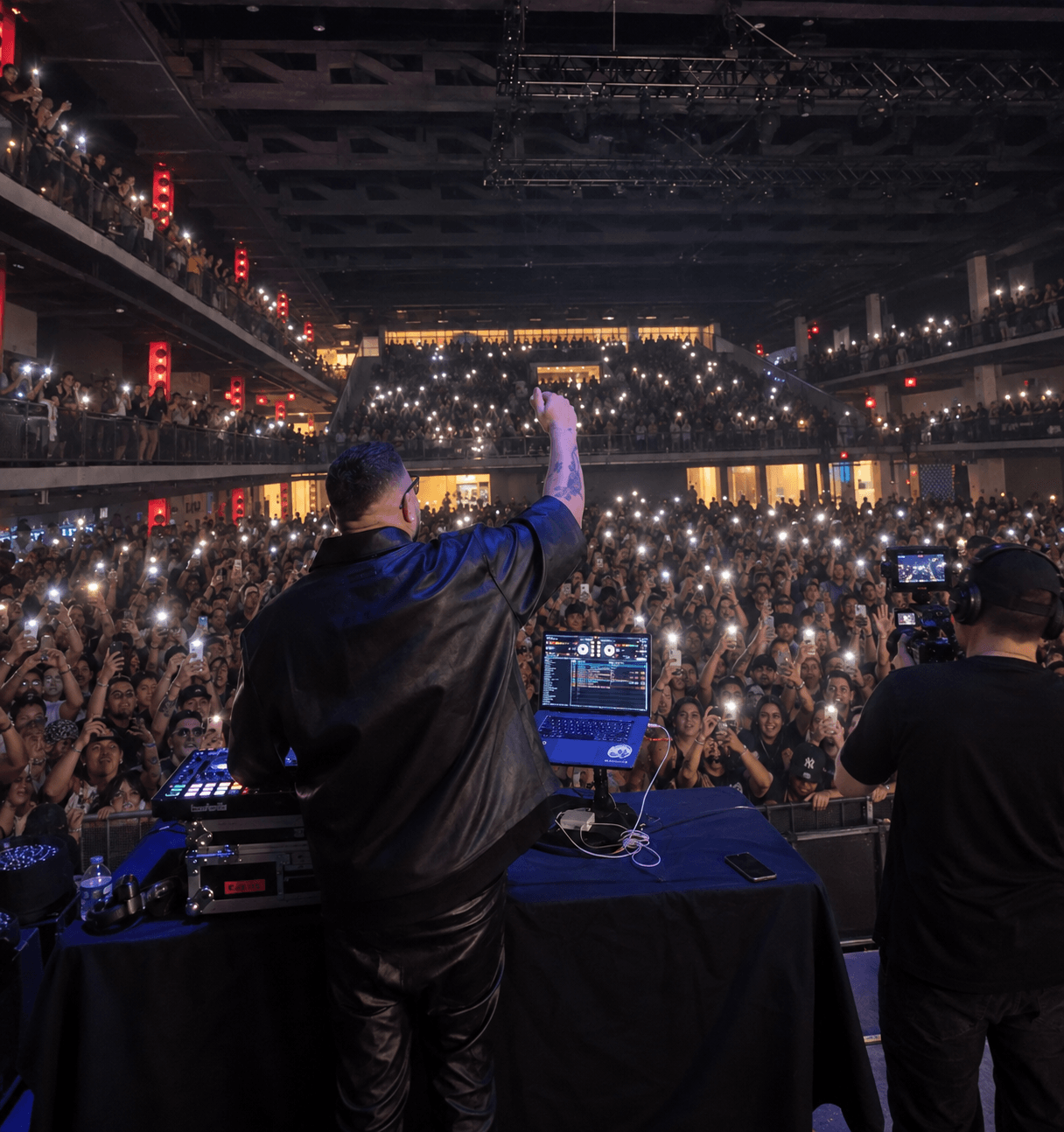 DJ performs on stage with crowd holding up lights in a large indoor venue, multiple levels, dark lighting, and equipment visible.