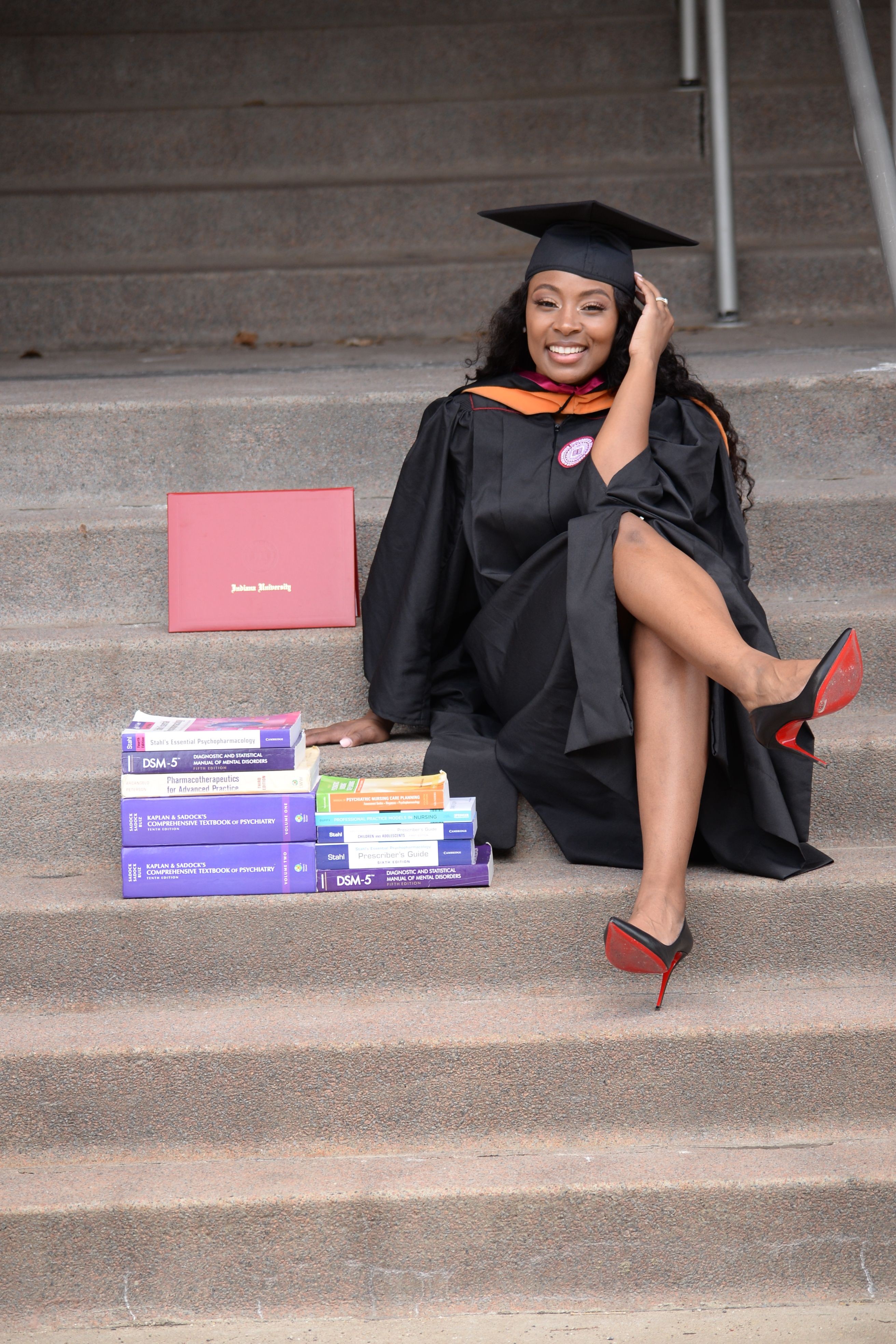 Person sitting on stairs with books and a pink box, wearing a black coat and red high heels, talking on phone.