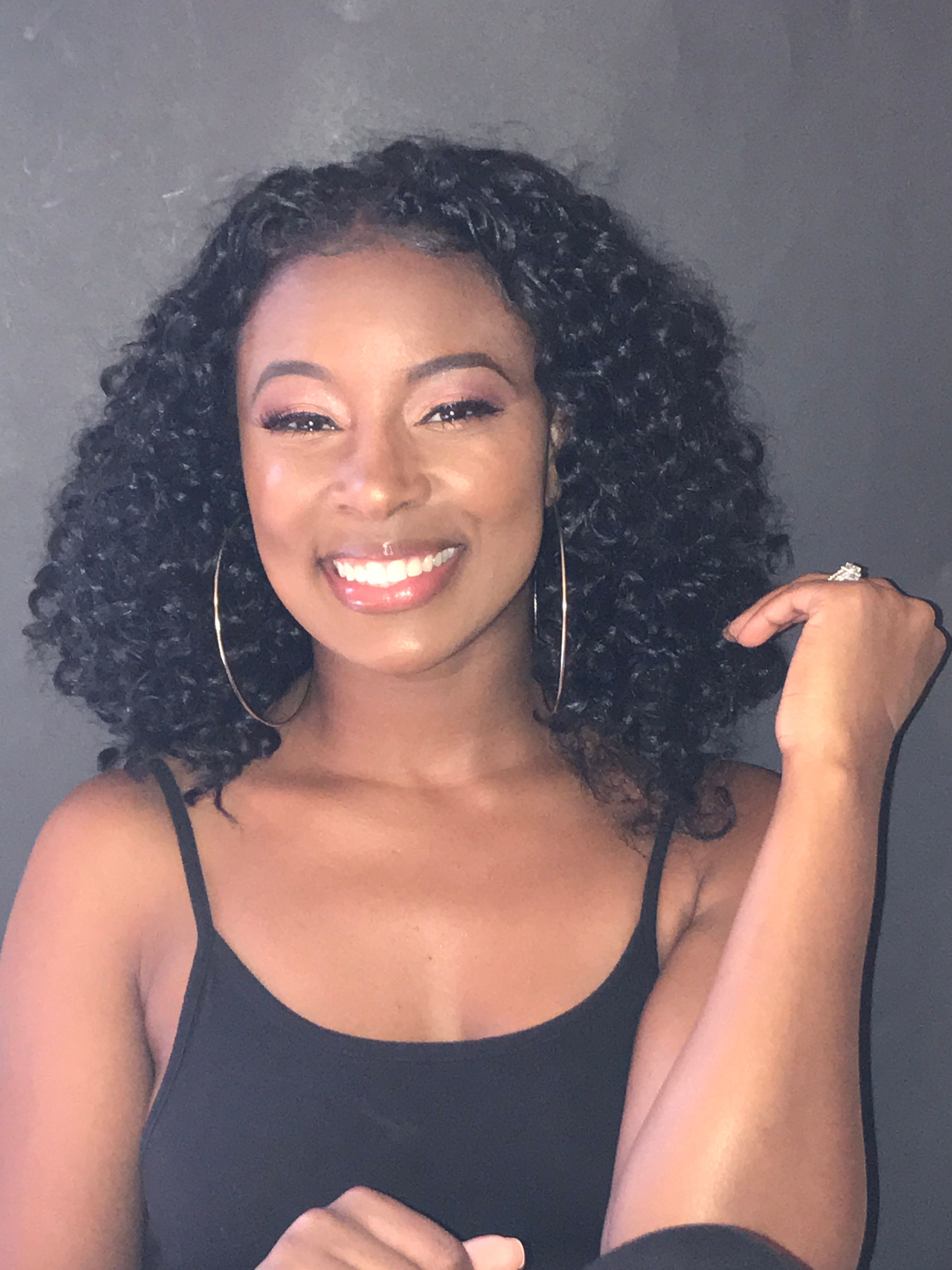Woman with curly hair smiling, wearing a black top, against a dark background.
