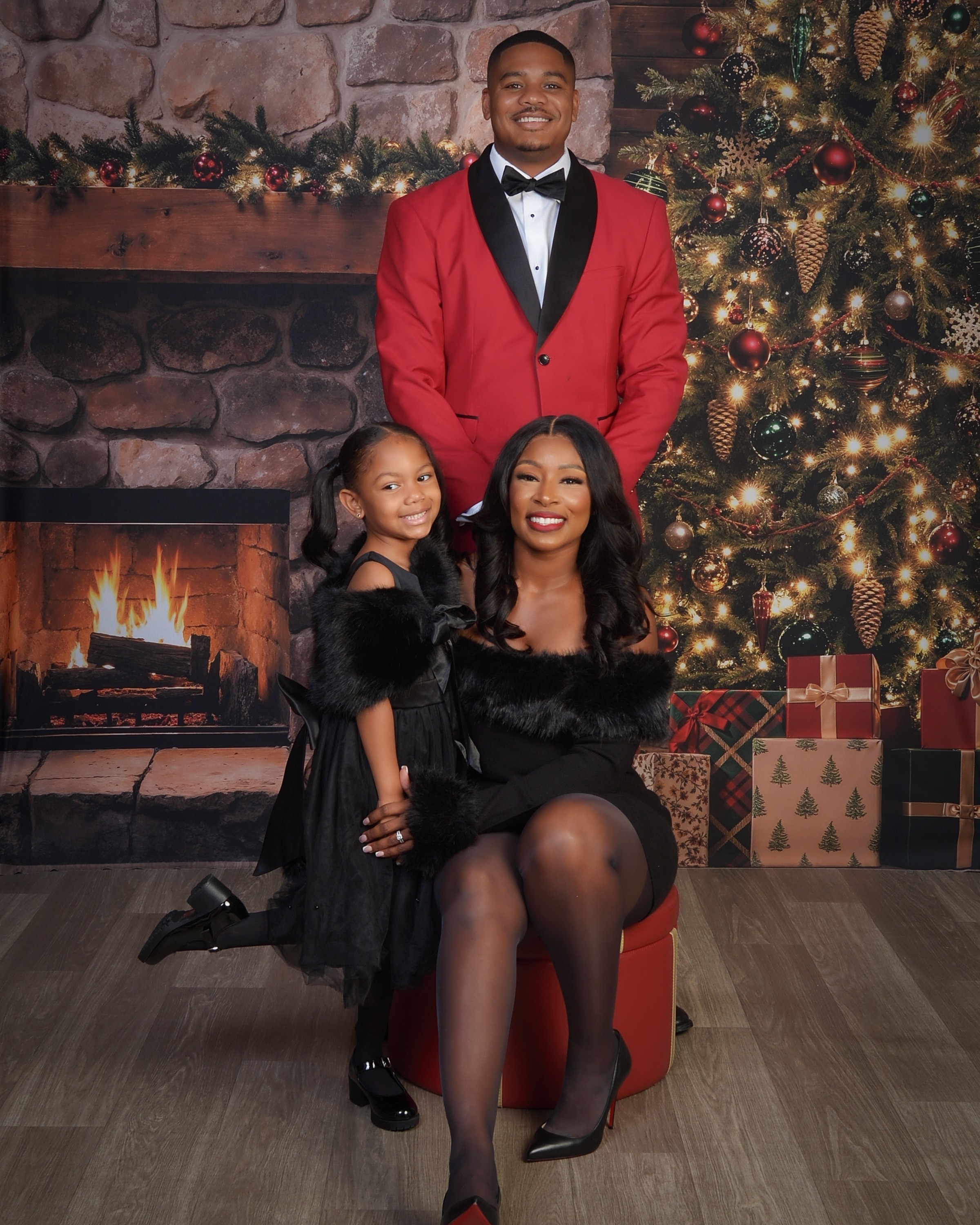 Three people in front of a decorated Christmas tree and fireplace, smiling, with wrapped presents nearby.