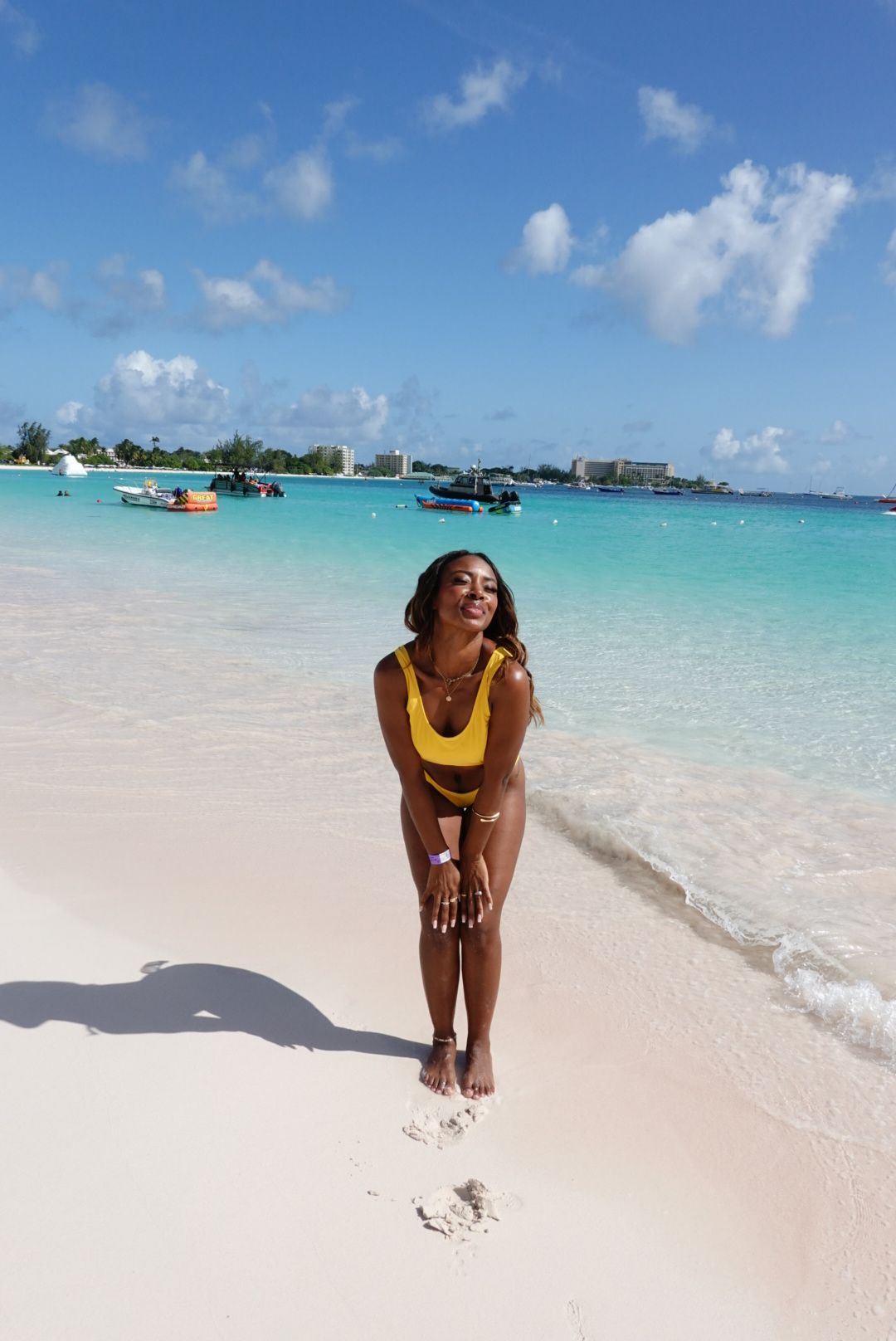 Woman in yellow swimsuit standing on sandy beach with turquoise water and boats in background under blue sky
