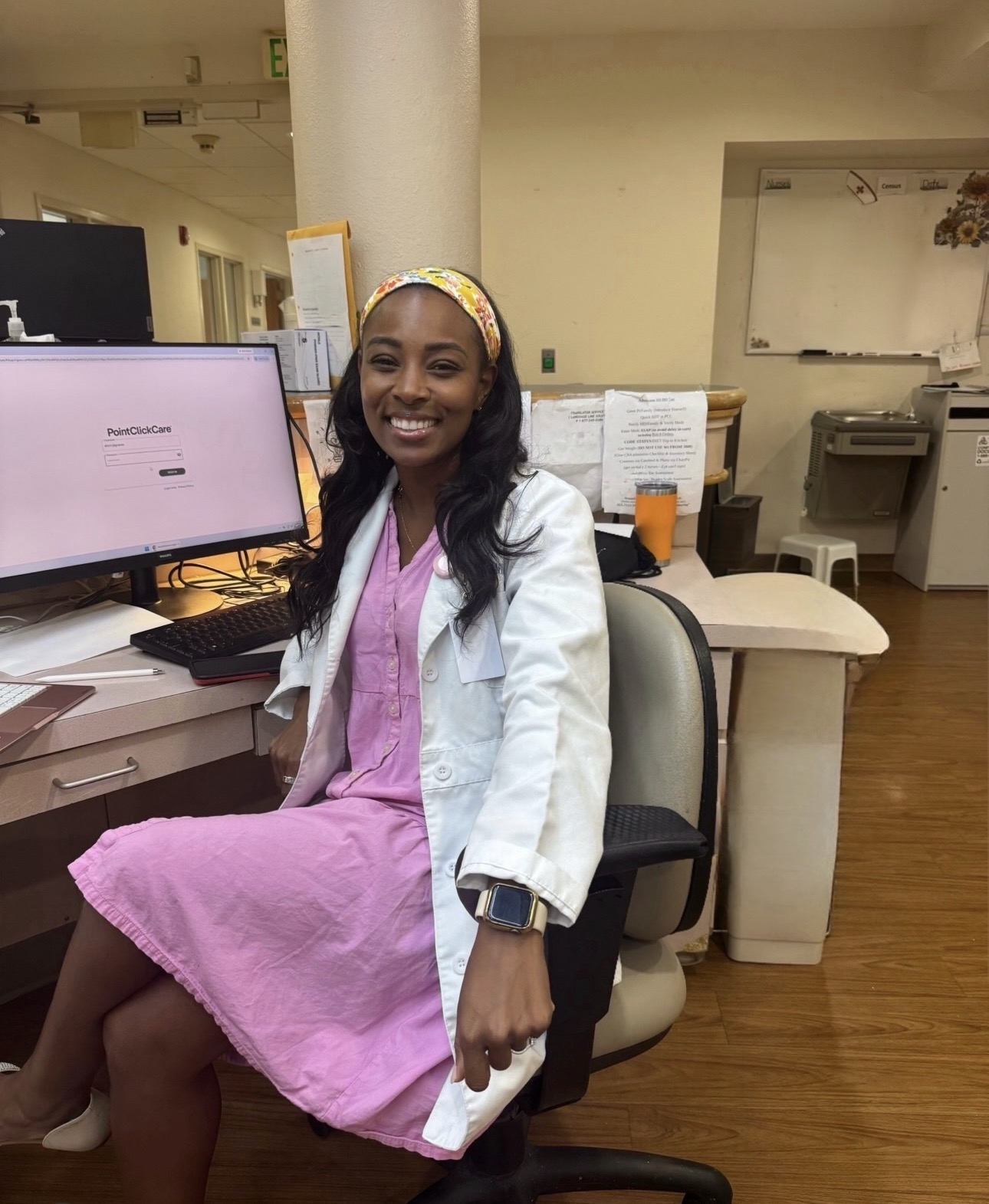 Smiling woman in pink dress and white coat sitting at a desk in an office or clinic, with computer monitor and papers.