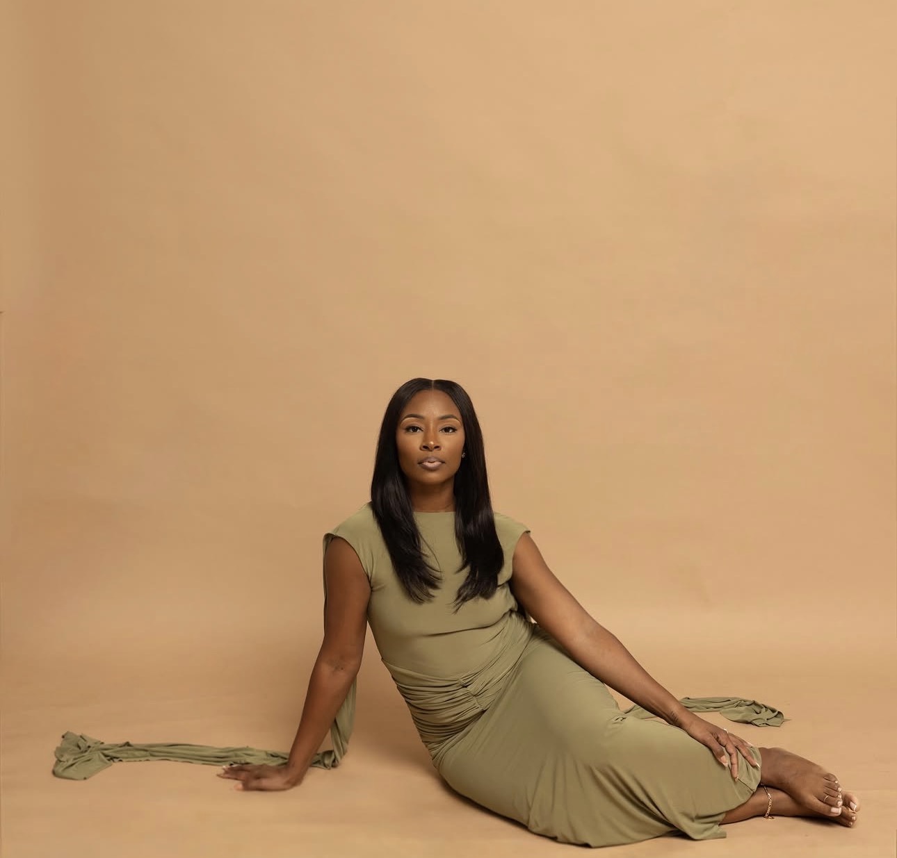 Woman in a long, olive-green dress sitting on the floor against a plain beige background.