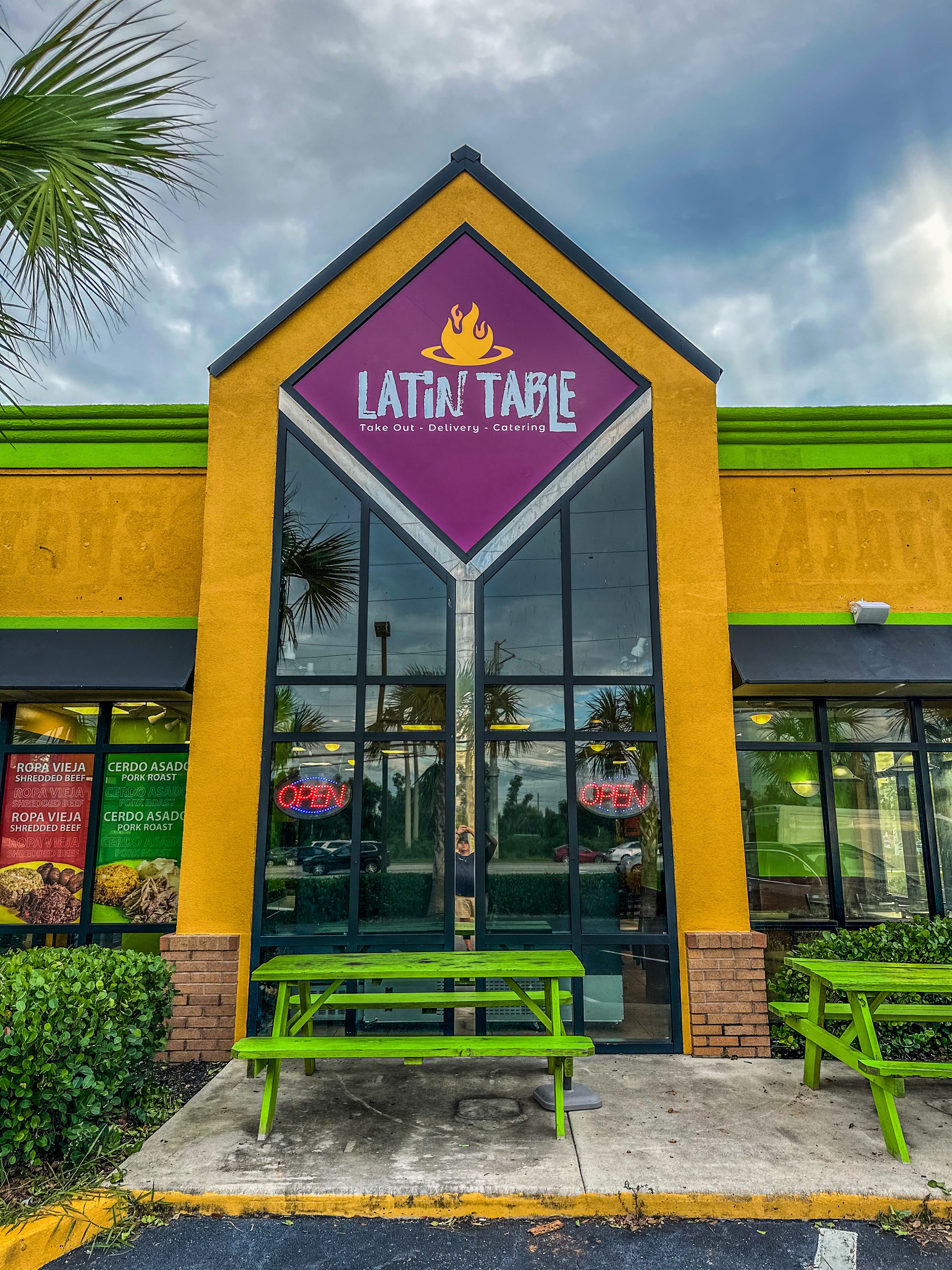 Front view of a restaurant with a purple sign reading 'Latin Table' and a yellow and green exterior, glass doors, and green benches.
