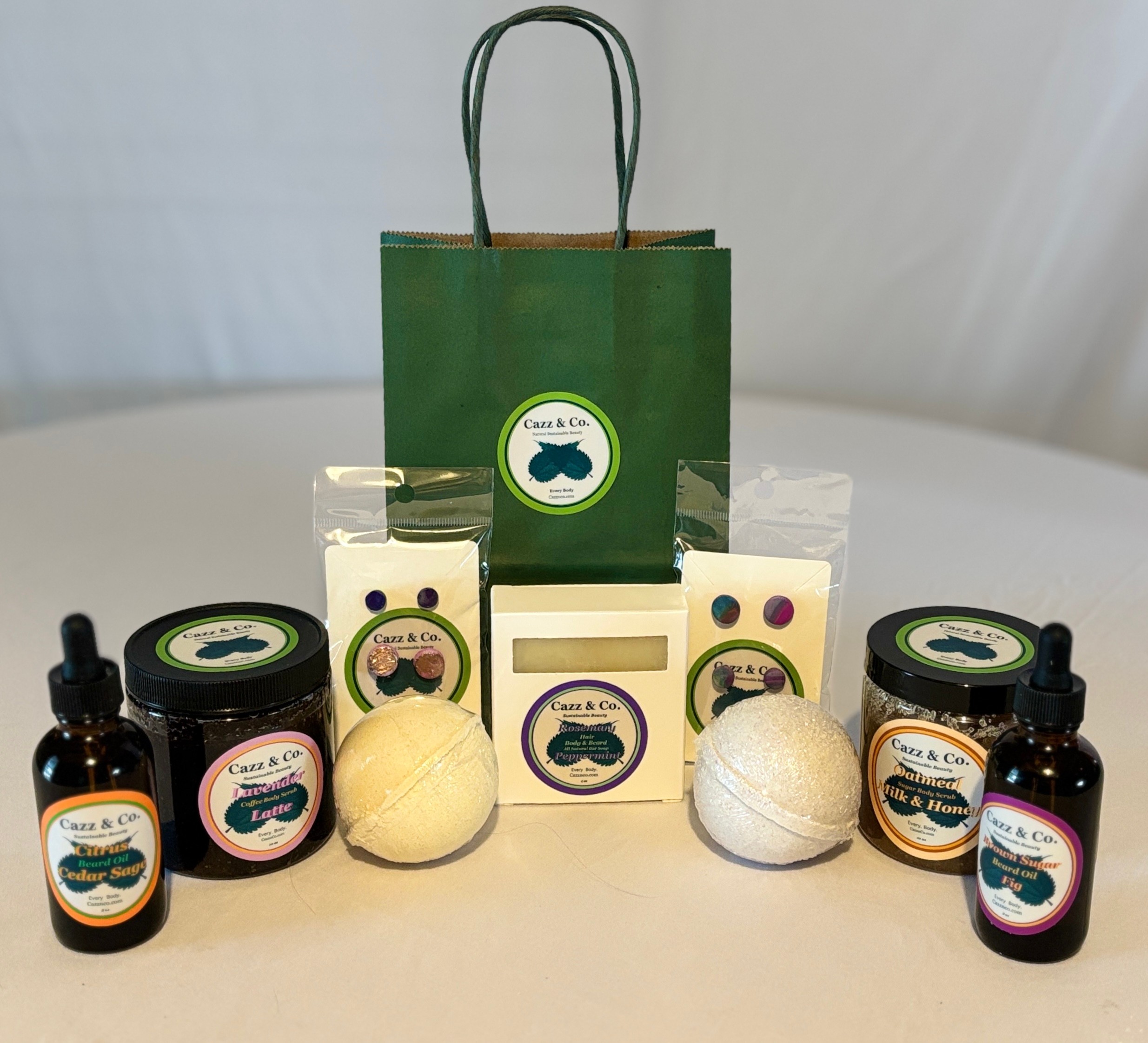 Green gift bag with handles, surrounded by skincare products, two bath bombs, and small bottles on a white surface.