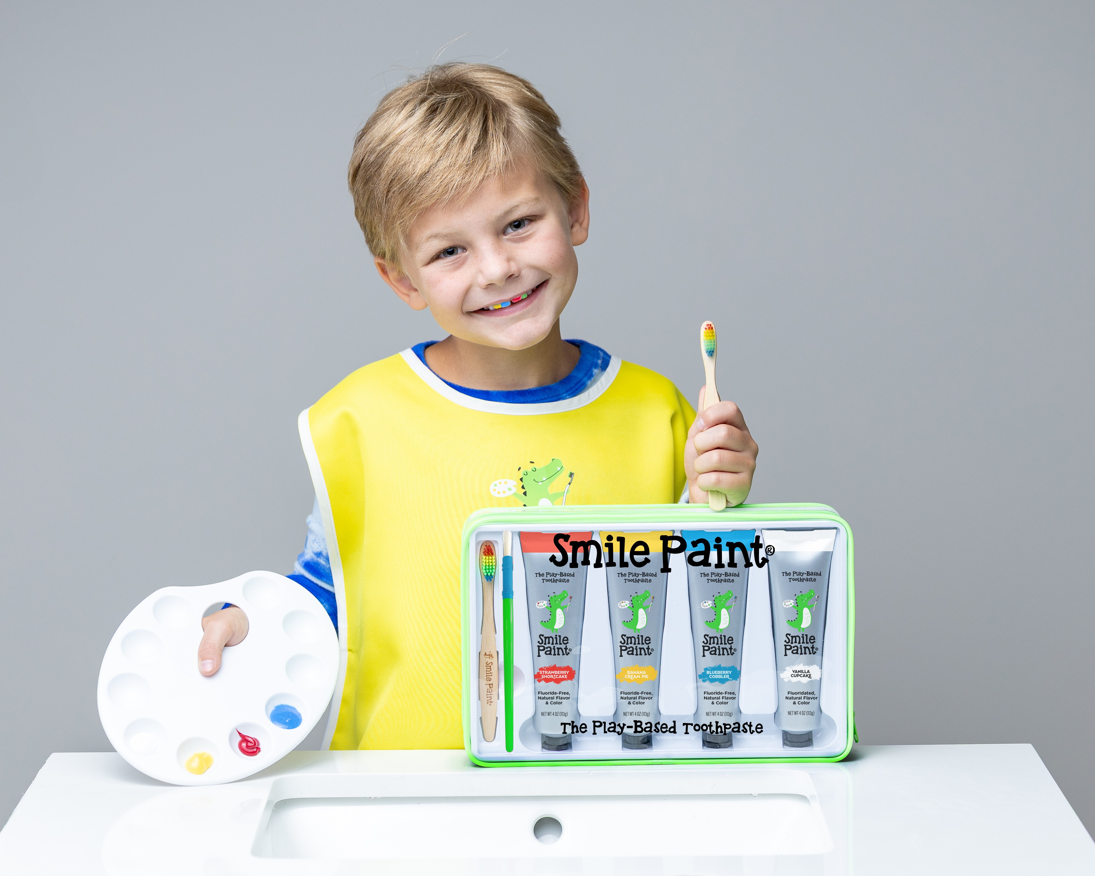 Child smiling holding a toothbrush, with a paint palette and a set of paint tubes on a table.