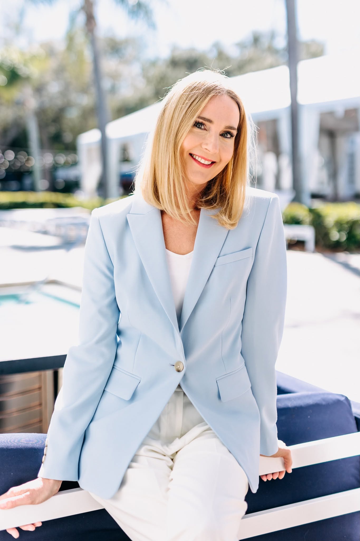 Woman with blonde hair smiling outdoors, wearing a light blue blazer and white top, with a blurred background of buildings and trees.