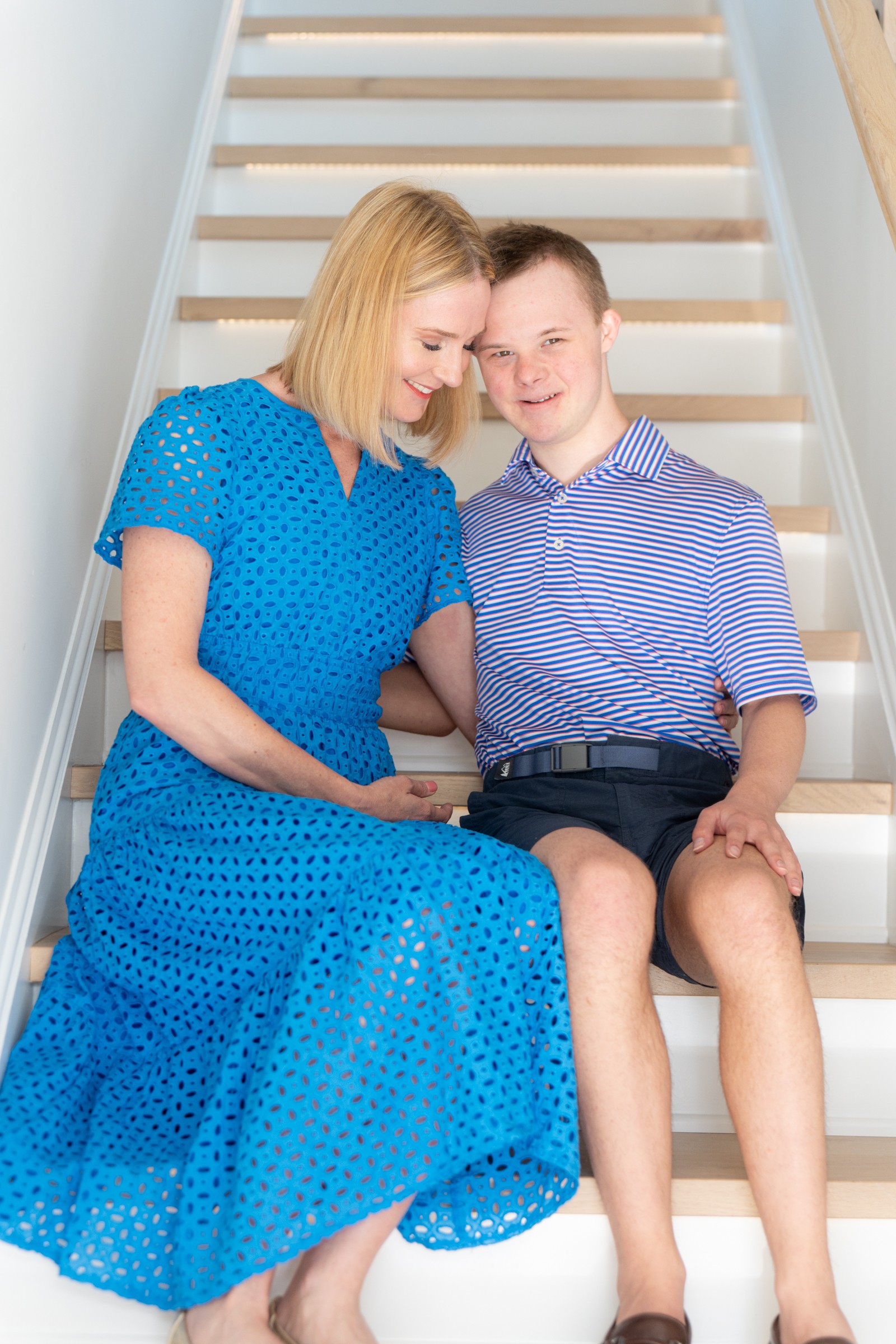 Woman and boy sitting on stairs, smiling, woman in blue dress, boy in striped shirt, close together.