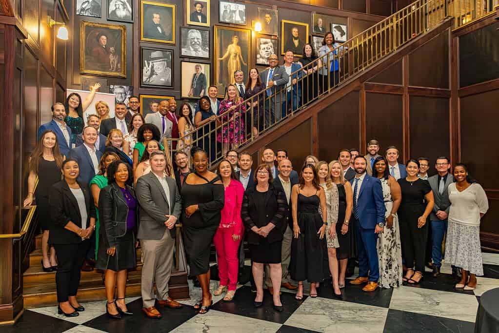 Group of people in formal attire standing in a room with framed portraits on the wall, some on stairs, smiling.