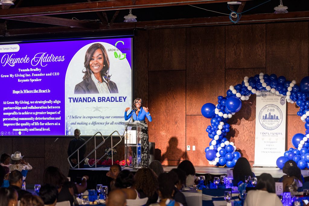 Woman speaking at a podium on stage with a large screen displaying her photo and information, decorated with blue and white balloons.